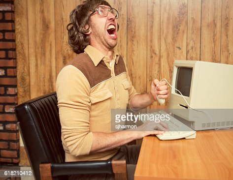 A retro looking funny angry computer office worker holding mouse and yelling in frustration.  Wood paneling office with vintage computer and keyboard on desk.